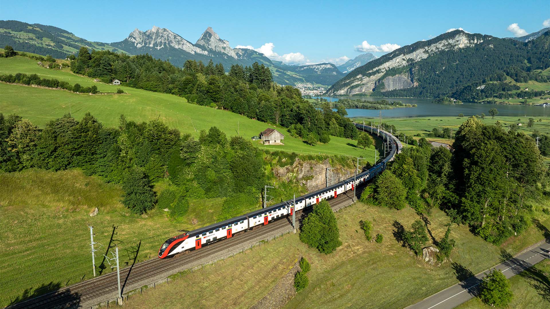 SBB-Doppelstockzug fährt auf Gleisen durch eine grüne Hügellandschaft mit Bergen und See im Hintergrund